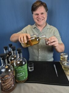 Distiller Matthew Mancuso stands at a bartop with a blue background, pouring amaro recipe ingredients into a small beaker and smiling at the camera